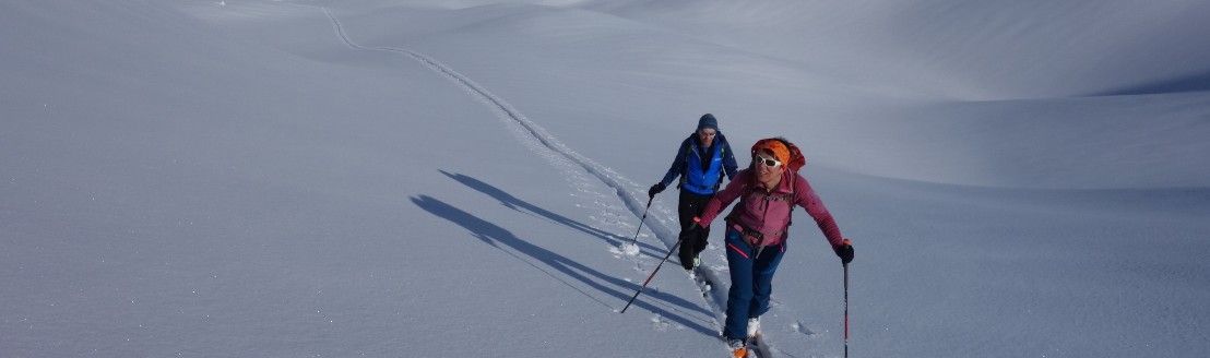 Eine Frau und ein Mann steigen auf einer Schneeflaeche Richtung Plueschhohe hoch